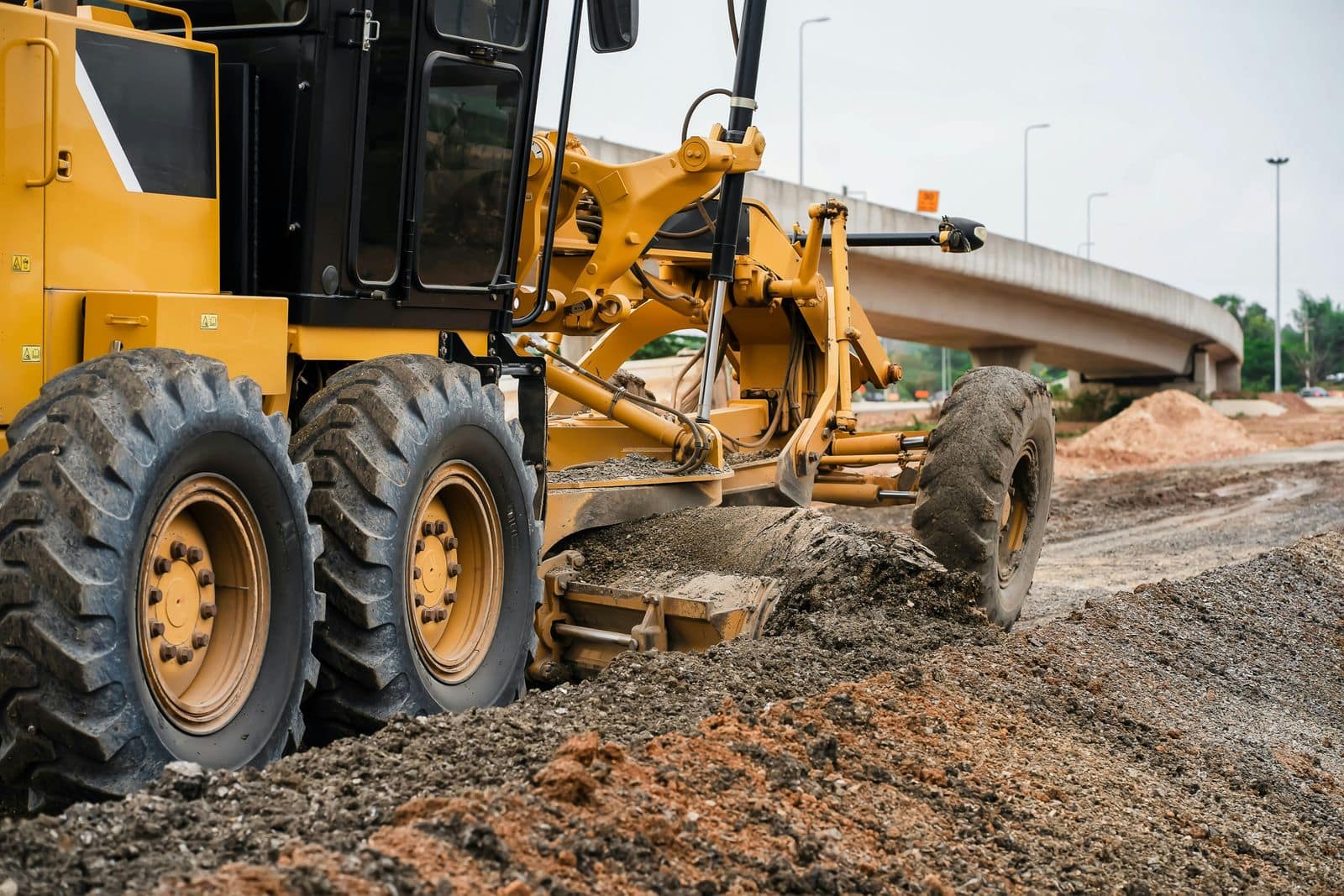 A motor grader spreading and levelling gravel base on a road under construction, with a highway overpass behind.